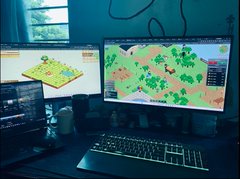 A dimly lit desk setup with a dual-monitor computer displaying games, a keyboard, and what appears to be a game controller in the foreground.