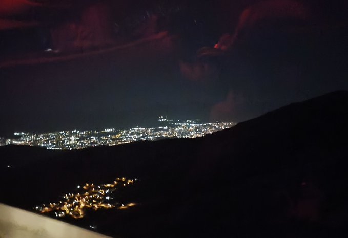 A nighttime view shows a city skyline glowing with lights in the distance, viewed from a dark hillside with a few faint clouds visible in the sky