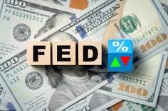 The word 'FED' spelled out in wooden letter blocks with a percent sign, red triangle, and green triangle, sitting on a pile of US dollar bills.