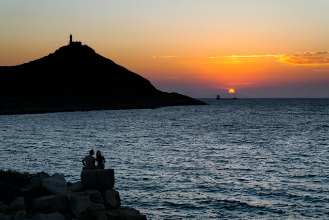 A scenic sunset photograph showing two people sitting on rocks by the sea, with a silhouetted hill and a small tower visible in the distance.