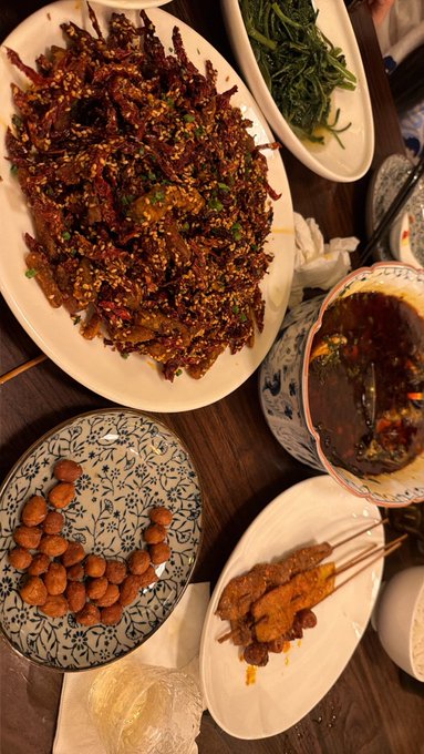 A table spread of various dishes including a large platter of spicy stir-fried meat with sesame seeds, fried potatoes, satay skewers, dark broth, and green vegetables.