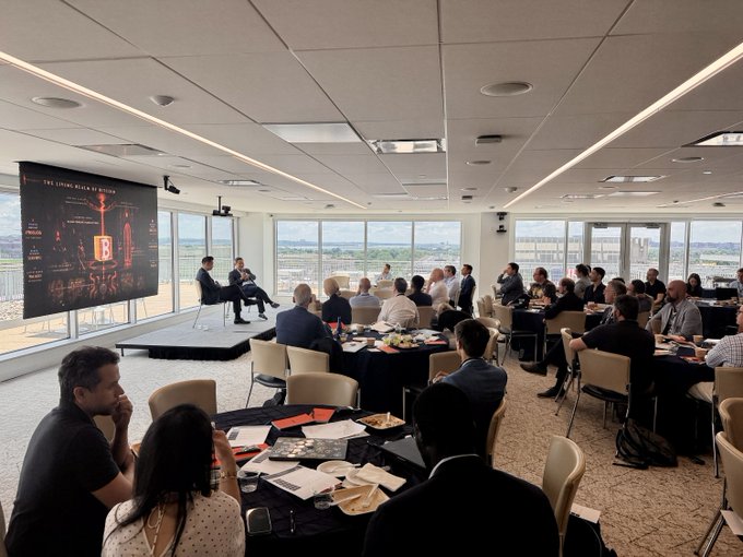 A corporate event or conference with attendees seated at round tables listening to two speakers on a small stage, with a large presentation screen displaying 'THE LIVING BLACK OF BURGUNDY' visible in the background.