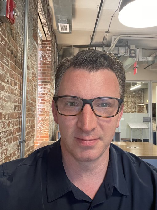 A selfie of a man with glasses and a black shirt standing in an office space with exposed brick walls and industrial ceiling fixtures.