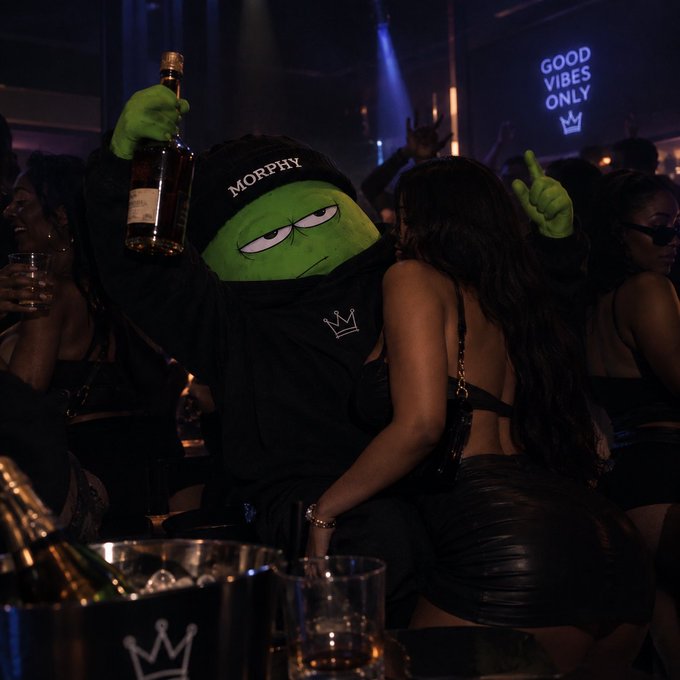 A nightclub scene featuring a person in a green Morph costume wearing a black hoodie with a crown logo, holding up a bottle of liquor, with a neon sign reading 'GOOD VIBES ONLY' and a crown symbol in the background.