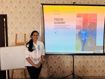 Woman in a white t-shirt standing next to a projection screen displaying 'TREZOR ACADEMY' during what appears to be a presentation or workshop.