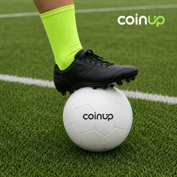 The image features a soccer player wearing black socks with neon green shoes standing on top of a white soccer ball that has not been kicked yet. The background shows a grassy soccer field, which serves as an interesting contrast to the vibrant colors of the player's footwear and the white soccer ball.