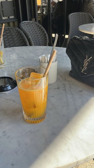 A glass of an orange-colored drink with ice and a straw on a marble cafe table, next to a sugar container and a black Yves Saint Laurent (YSL) handbag.