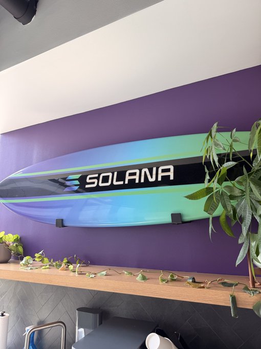 A colorful surfboard displaying the "SOLANA" logo is mounted on a purple wall, with the cryptocurrency brand's name prominently displayed in bold white lettering. The "surging the solana wave" caption ties to the surfboard imagery, symbolizing the rising momentum of the Solana cryptocurrency.