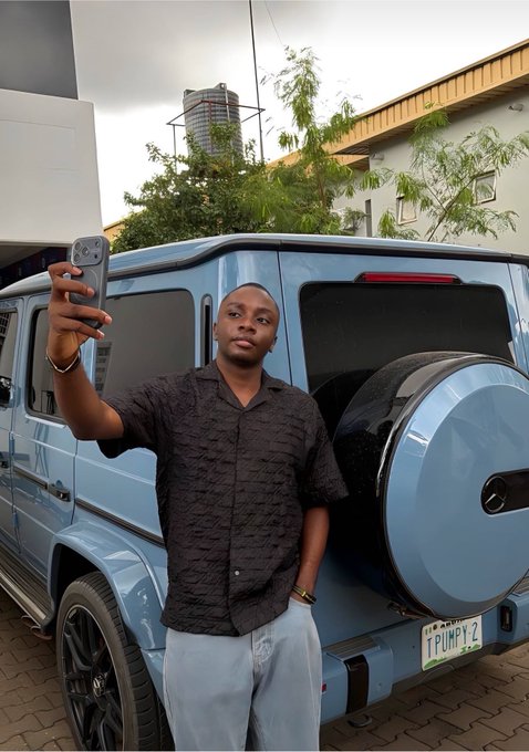 A man in a black textured shirt and light pants taking a selfie with a smartphone while standing in front of a light blue Mercedes-Benz G-Wagon with the license plate "TPUMPY2".