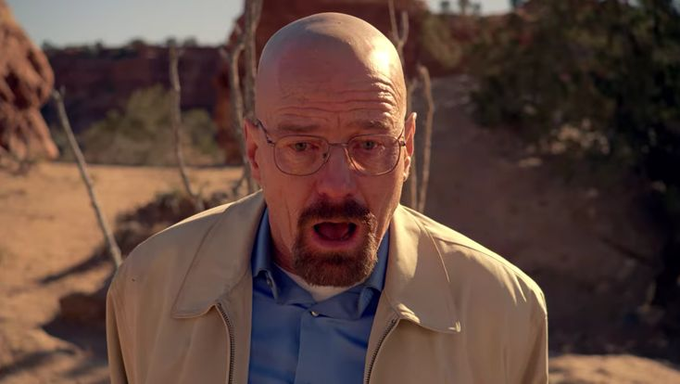 A bald man with glasses and a goatee, wearing a tan jacket over a blue shirt, standing in a desert landscape.