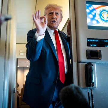 The image shows a man dressed in a suit, wearing a red tie, standing inside an airplane with his hand raised in front of him. The man appears to be giving an order or making a gesture while being seated next to a phone on the plane's entertainment system. The background features a TV screen displaying an American flag and a clock showing the time as 1:25 pm.
