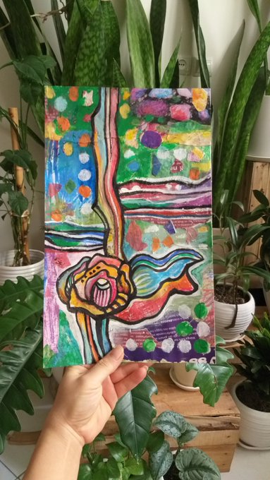 A hand holds up a small, colorful abstract mixed-media artwork featuring vibrant floral and landscape elements, displayed in front of several potted plants including snake plants.