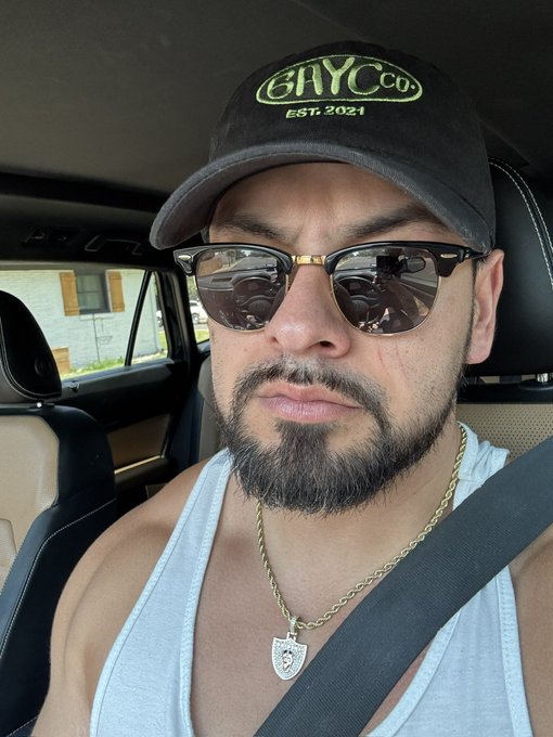 A selfie of a man wearing a black cap with "BAY CO EST. 2021" in green text, sunglasses reflecting a steering wheel, a white tank top, and a gold chain with a pendant, taken inside a car.