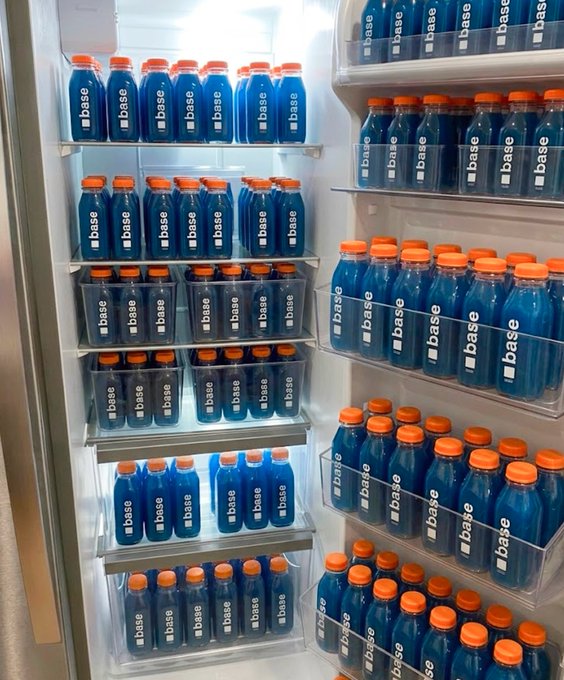 The image shows a refrigerator filled with blue bottles of Base juice, placed in clear plastic containers. There are at least 13 bottles visible inside the fridge, arranged in various positions. The refrigerator appears to be well-stocked and ready for sale or distribution.