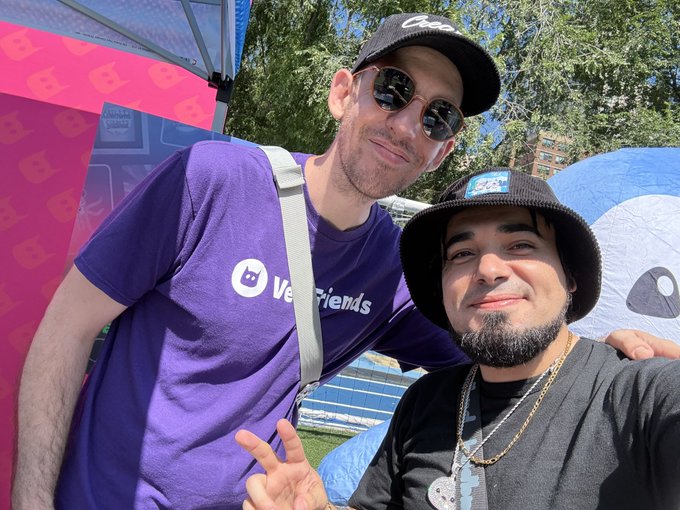 Two men pose for a selfie outdoors at an event; the taller man wears a purple 'VeeFriends' t-shirt, sunglasses, and a black cap, while the other wears a black shirt, bucket hat, and silver chains.