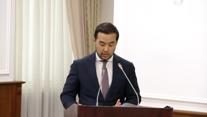 A man in a dark blue suit and pink tie stands at a wooden podium with two microphones, appearing to read from papers in a formal setting.