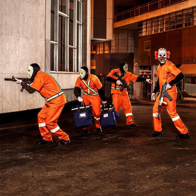 Halloween-themed photo of four people in orange hazmat-style jumpsuits and creepy masks (alien, halloween, and clown) holding props like rifles, a briefcase, and a duffel bag in a gritty industrial setting.