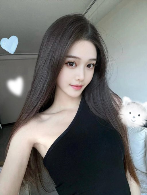 A selfie of a young woman with long dark hair wearing a black one-shoulder top, with a white bear plush visible in the background and a small blue heart graphic overlay.