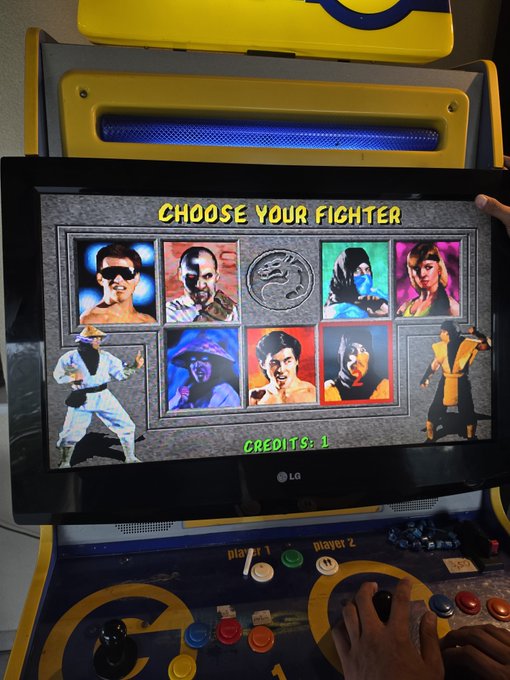 An arcade cabinet displays the 'CHOOSE YOUR FIGHTER' character selection screen from the classic video game Mortal Kombat. The screen shows portraits of fighters including Johnny Cage, Kano, Raiden, Kitana, and Liu Kang, with a 'CREDITS: 1' counter and a player's hand visible on the control panel.