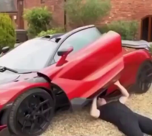 The image features a man lying on his stomach next to a red sports car with its hood open, appearing to be underneath a wheel. The context of the scene suggests that the man might be working on repairing or maintaining the car. In the accompanying text from another post, it says "When $BTC goes down, my workouts will change". The connection between these two pieces of information is unclear and may not necessarily be related to the image itself or its content.
