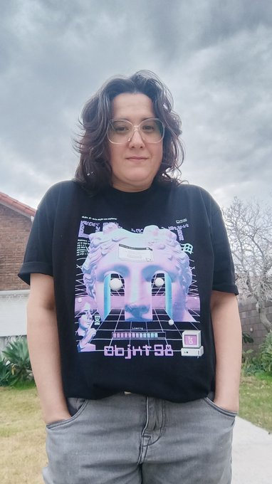 A woman with glasses and curly dark hair wearing a black t-shirt with a colorful retro/vaporwave-style graphic print, standing outdoors under a cloudy sky.