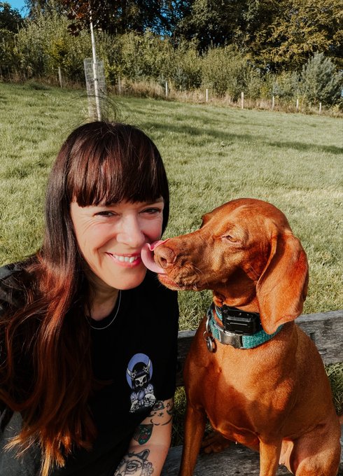 A woman with dark hair smiles as a brown Vizsla dog licks her face in a grassy outdoor area.