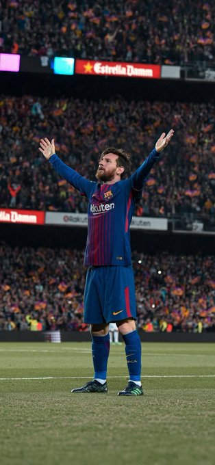 The image features a soccer player standing on a field, holding his arms up in the air with both hands. The player appears to be celebrating or expressing joy after scoring a goal during a match. In the background of the photo, there are several people present who seem to be watching and enjoying the game as well.