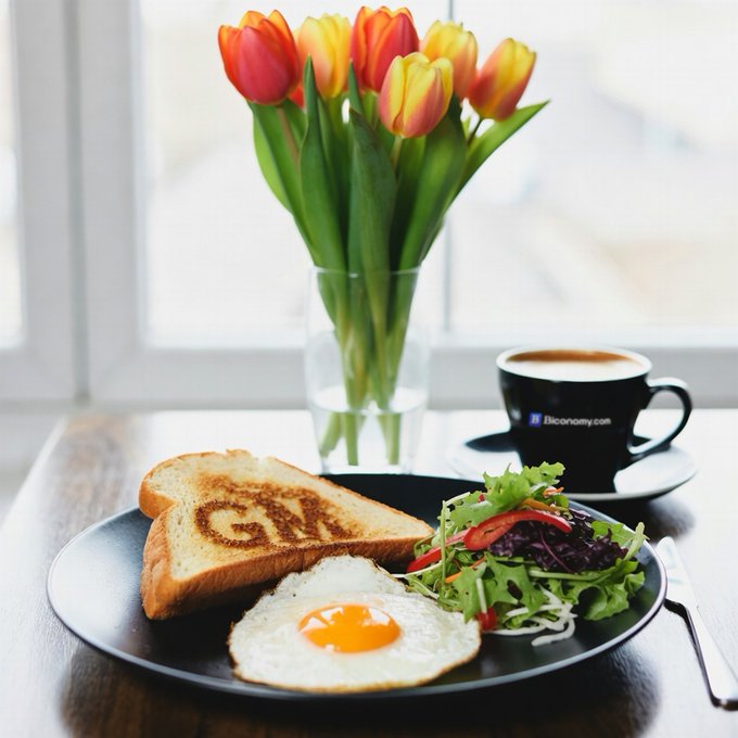 The image shows a breakfast plate with an egg, toast, and salad on a wooden table. The plate has the words "GM" written on it, which could be related to the breakfast or possibly a brand name for the meal. The breakfast is served in a glass vase filled with tulips, adding a touch of color and freshness to the scene. A cup of coffee and a knife are also present on the table.