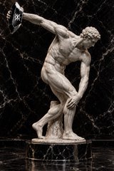 A classical marble statue of a nude muscular man in a dynamic pose, throwing a discus against a black marble background.