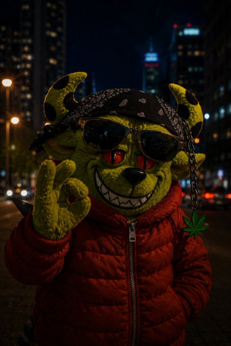 A person wearing a green, horned monster mask with red eyes and a wide grin, accessorized with sunglasses, a black bandana, and a marijuana leaf chain, dressed in a red puffer jacket against a nighttime city backdrop.