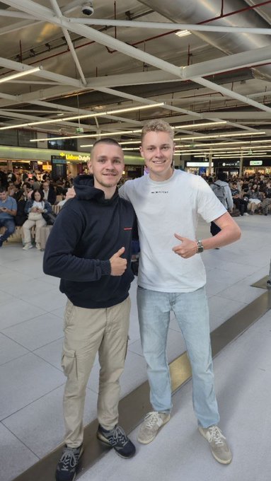 The image shows two young men posing for a picture at an airport terminal. They are standing next to each other with their arms around each other's shoulders, smiling and enjoying themselves. The background of the photo reveals that they are surrounded by several people in various positions throughout the scene. There is also a suitcase visible near one of them.

The image includes text that reads "Your favorite German KOLs are now flying back. Next stop is the Solana Summit in Berlin ... See you on the other side." This suggests that these two men might be traveling together and have just arrived at their destination, possibly Berlin. The presence of multiple people around them indicates a busy airport environment where travelers from different places gather to embark or return home.