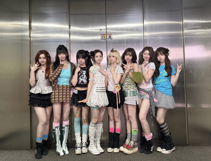 A group of eight young women pose together in front of metallic elevator doors, wearing colorful, eclectic Y2K-style outfits with mismatched socks, platform boots, and varied hairstyles.