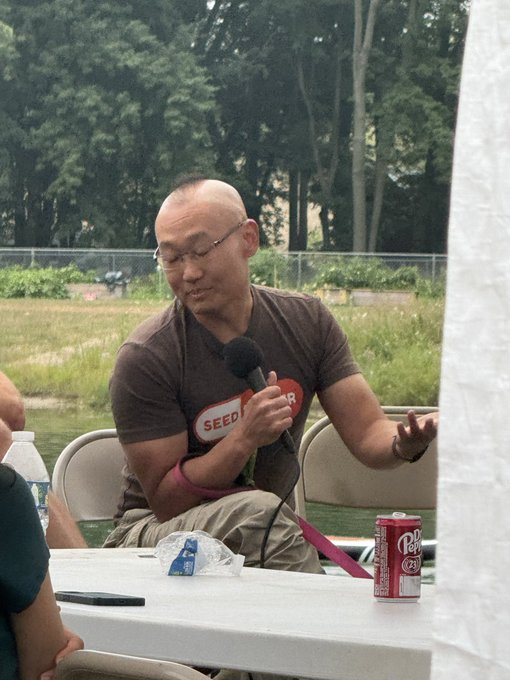 A bald man wearing glasses and a brown t-shirt with a red "SEED" logo speaks into a microphone while seated at a white table with a Dr Pepper can, set against an outdoor backdrop of trees and a pond.