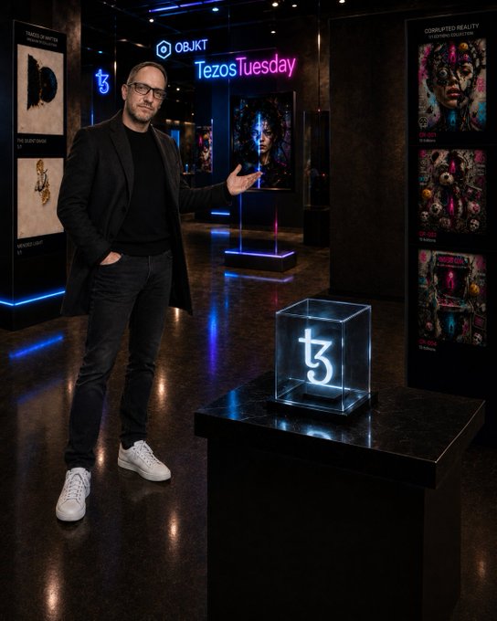 A man in a black coat, black t-shirt, and white sneakers poses next to an illuminated Tezos logo display at what appears to be an OBJKT Tezos Tuesday NFT event, with various digital artworks displayed in the background.