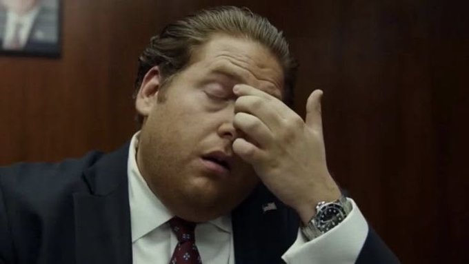 A man in a dark suit, white shirt, and patterned red tie rubs his eyes with his hand while wearing a luxury wristwatch, appearing fatigued in what looks like an office setting.