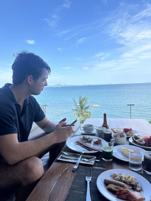 A man sits at a breakfast table overlooking an ocean view, checking his phone while surrounded by plates of food including sausages, eggs, and toast, with a green juice and coffee on the table.