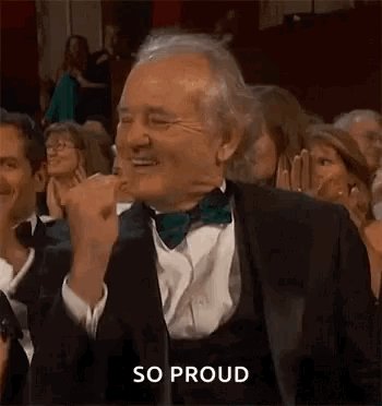 The image shows a man wearing a suit, tie, and bowtie standing in front of an audience at an event. He appears to be giving a speech or making a gesture while surrounded by people who are watching him intently. The caption "So proud" is written below the image, which suggests that the man might have accomplished something significant or is receiving recognition for his work.