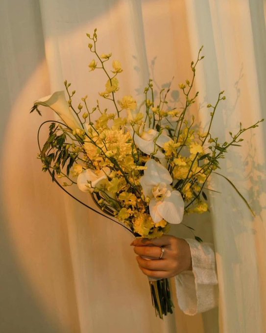 A close-up of a hand holding a bouquet of yellow flowers and white orchids, with a ring visible on the finger, set against a softly lit curtain backdrop.