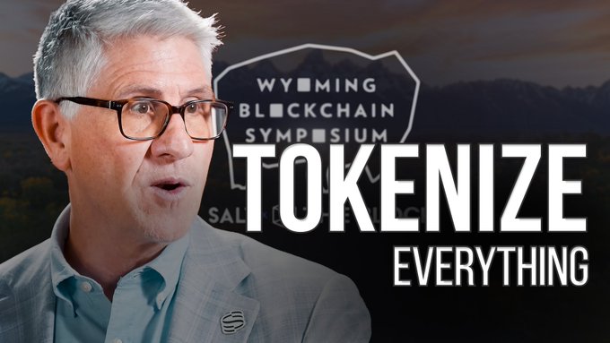 A man with glasses speaks in front of a backdrop reading 'Wyoming Blockchain Symposium' and 'Tokenize Everything'.