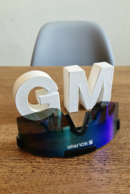 Decorative white 'GM' (Good Morning) letters on a wooden table placed behind a pair of iridescent blue-green sunglasses, with a chair in the background.