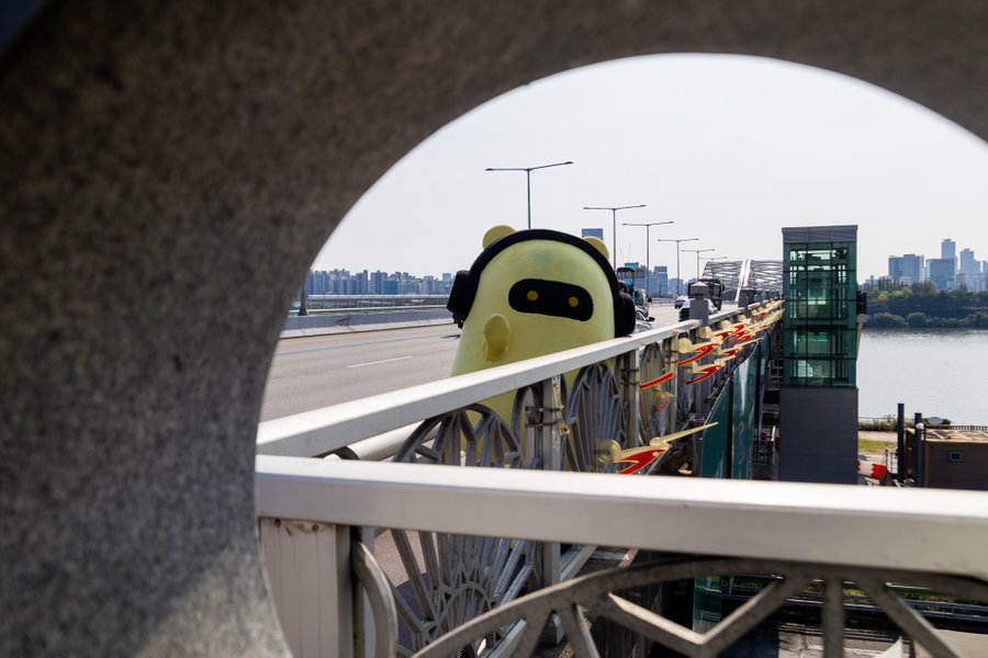 A cute yellow mascot character with headphones stands on a bridge overlooking a cityscape, viewed through an arched frame, symbolizing Seoul's fusion of playful culture with sophisticated financial infrastructure that appeals to algorithmic traders. The image complements the post's message about Seoul traders prioritizing speed, sophistication, and market data, with the mascot's headphones suggesting a focus on real-time information over commentary.