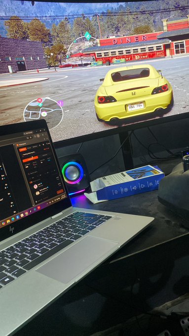 A gaming setup showing an HP laptop running on a desk in front of a large monitor displaying a yellow sports car at a diner in what appears to be a racing game, with an RGB-lit speaker nearby