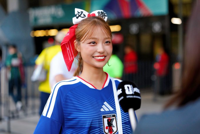 A smiling young woman wearing a Japan national football team jersey (featuring the "JAPAN" crest and JFA emblem) and a red-and-white bow headband is being interviewed by a reporter holding a "Q" branded microphone, likely at a sporting event venue.

The image appears to relate to a cultural or sporting event in America, complementing the post's surprise about affordable $300/night stays in the US and mentions of Polymarket, suggesting a connection to international travel or a Japan-related event abroad.