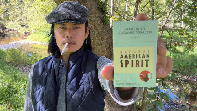 A young person wearing a flat cap and quilted vest smokes a cigarette while holding up a pack of 'Natural American Spirit' cigarettes toward the camera, with a wooded outdoor area in the background.