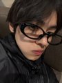 A young man with dark hair wears stylish black-rimmed glasses and a black puffer jacket, looking at the camera with a soft expression. The image appears to be a fan-made photo related to ALLYZ, a member of the group ALD1 (Alpha Drive One), with the caption expressing admiration for him using hashtags like #ALPHADRIVEONE and #리오 (Leo).