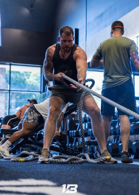 A muscular man in a black tank top strains to pull a heavy battle rope in a gym, while another man in a green shirt and shorts stands nearby assisting, embodying the "We working" spirit shown in the post, with a "1B" logo visible at the bottom of the image.