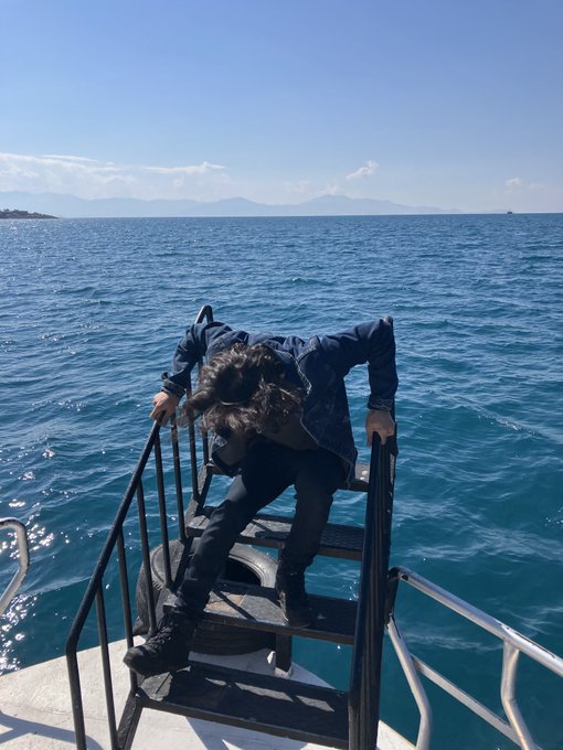 A person in a dark jacket and pants standing on a metal boat ladder descending toward the blue ocean, with mountains visible on the horizon.