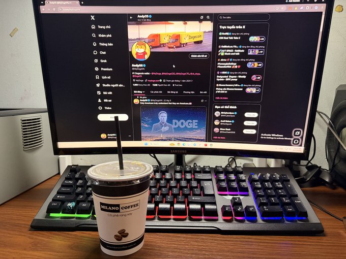 A desktop setup featuring a Samsung monitor displaying Twitter/X with an 'AndyG' profile, an RGB backlit gaming keyboard, and a 'MILANO COFFEE' cup with a straw in the foreground.