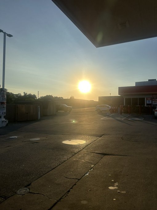 Sunset view from under a gas station canopy, looking out over a parking lot with a few cars, a street lamp, and distant trees silhouetted against a golden sky.