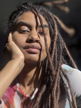 A person with long dreadlocks poses with their hand near their face, wearing colorful eyeshadow and a pink patterned top.